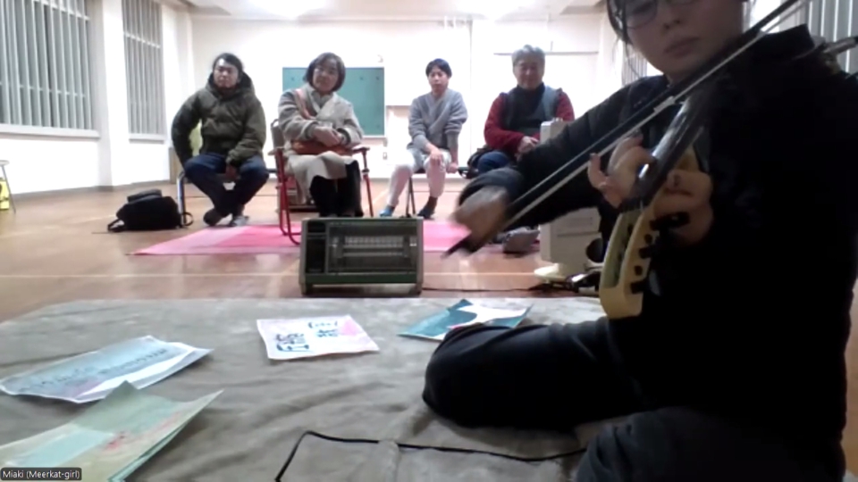 one of the artists, miaki, sits crosslegged playing the Cantini, a black MIDI violin, with a bow. Four audience members look on behind her.