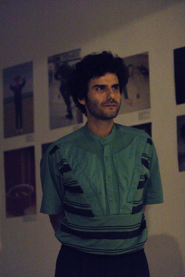 the artist looks on, standing, hands behind his back. He is wearing dark trousers and a green top with vertical buttons going down two thirds of the shirt. Black horizontal stripes run along the bottom and the sides of it.