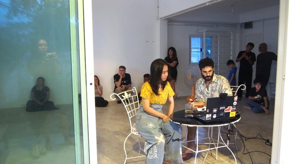 view of the artists seated at a small table with a glass of rum and a laptop on it. stephan e perez holds a tabacco pipe. The artists are seated facing the door with their backs to the audience, who are standing around the inside of the room